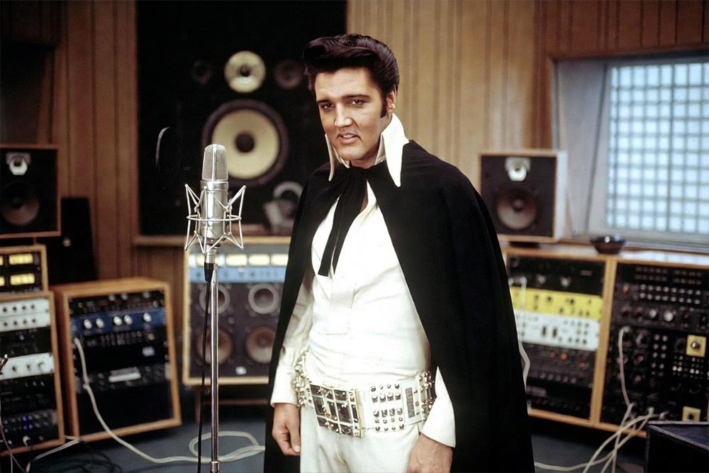 Elvis Presley w białym stroju i czarnej pelerynie przy mikrofonie w studiu nagraniowym