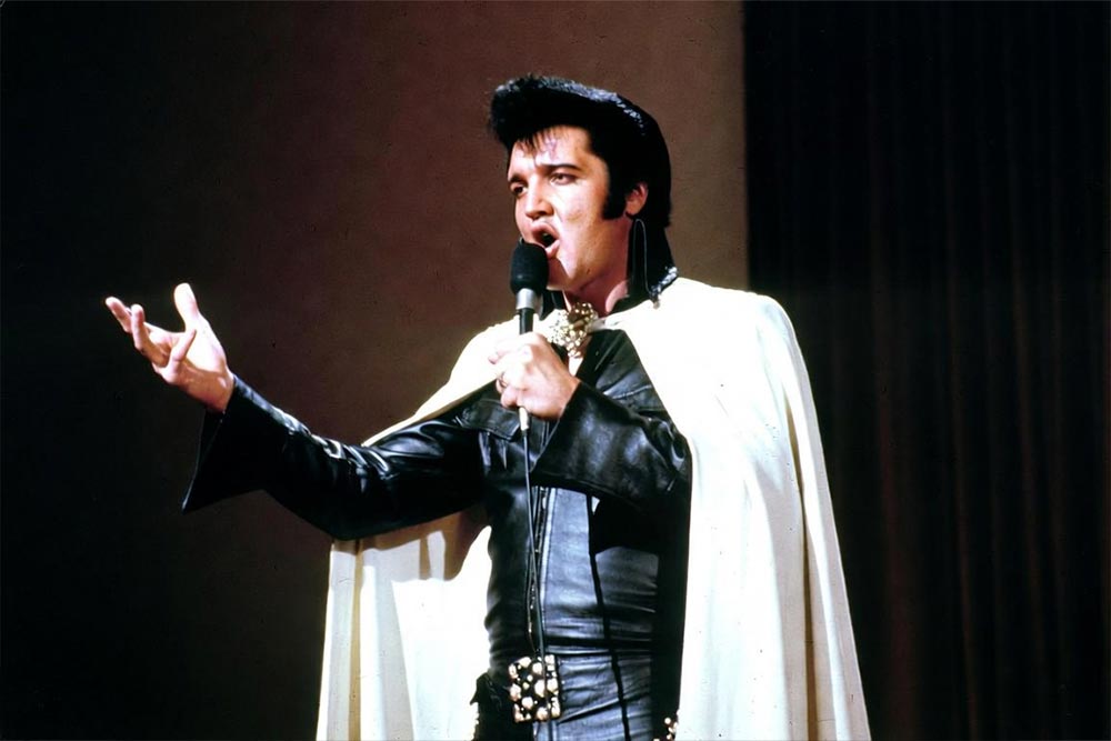 Elvis Presley śpiewający na scenie w czarnym skórzanym stroju i białej pelerynie
