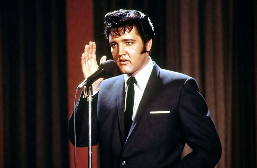 Elvis Presley w granatowym garniturze śpiewający przy mikrofonie na tle czerwonej kurtyny